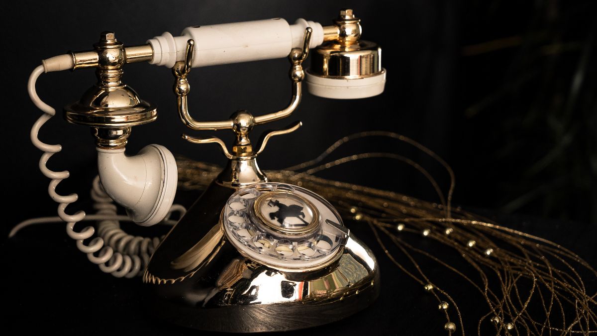 Retro telefón s číselníkom, zlaté detaily a biely sluchátko na tmavom pozadí.
