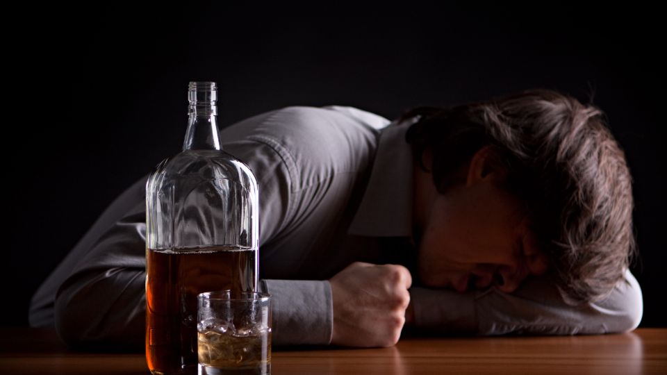Muž opretý o stôl s hlavou sklonenou, vedľa neho fľaša alkoholu a pohár.