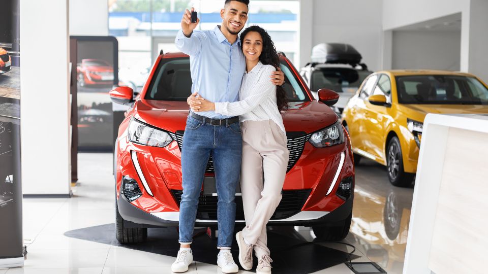 Šťastný pár stojí pred červeným autom v autosalóne, usmievajú sa a držia kľúče.