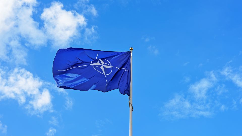 Modrá vlajka NATO s bielym symbolom na pozadí modrej oblohy.