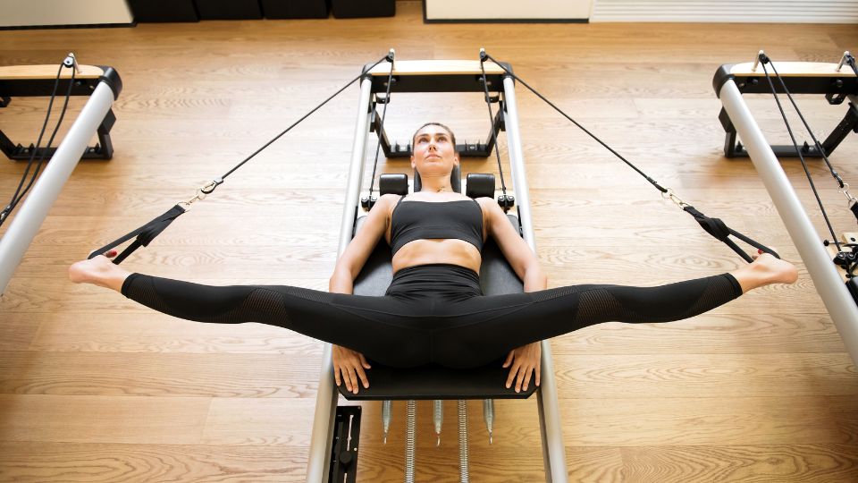 Žena cvičí na pilates stroji s rozkročenými nohami, zameraná na flexibilitu.