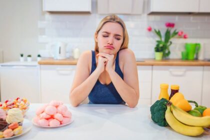 Žena sedí za stolom, zamyslená medzi nezdravými a zdravými jedlami.