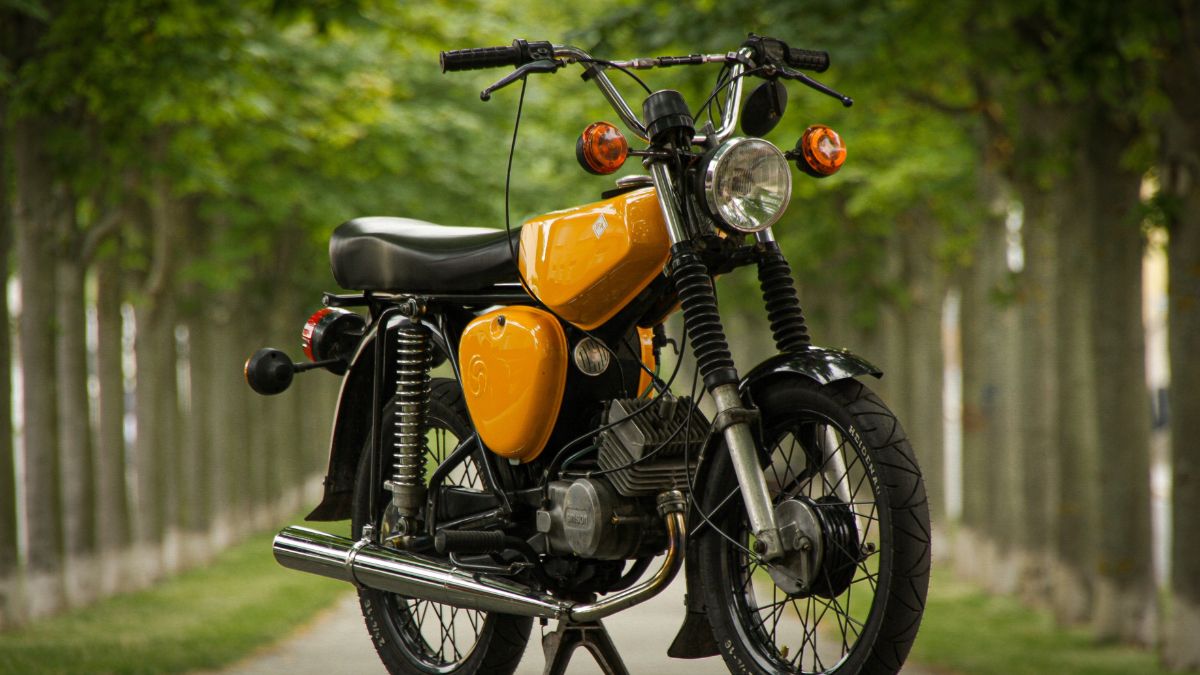 Motocykel Simson s oranžovou nádržou a čiernym rámom na ceste medzi stromami.