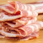 Usporiadané plátky slaniny na drevenej doske, s viditeľnou mastnou textúrou.
