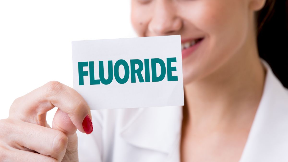 Žena v bielom plášti drží kartičku s nápisom 'FLUORIDE'.