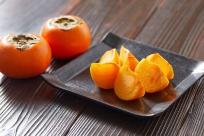 Dva celé hurmi kaki a plátky tomelu na čiernom tanieri.