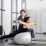 Žena cvičí na gymnastickej lopte s činkami, zatiaľ čo ju inštruktorka pozoruje.