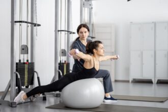 Žena cvičí na gymnastickej lopte s činkami, zatiaľ čo ju inštruktorka pozoruje.