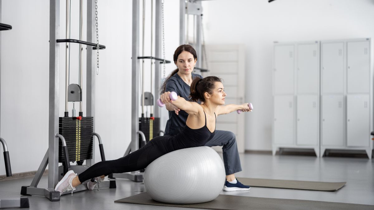 Žena cvičí na gymnastickej lopte s činkami, zatiaľ čo ju inštruktorka pozoruje.