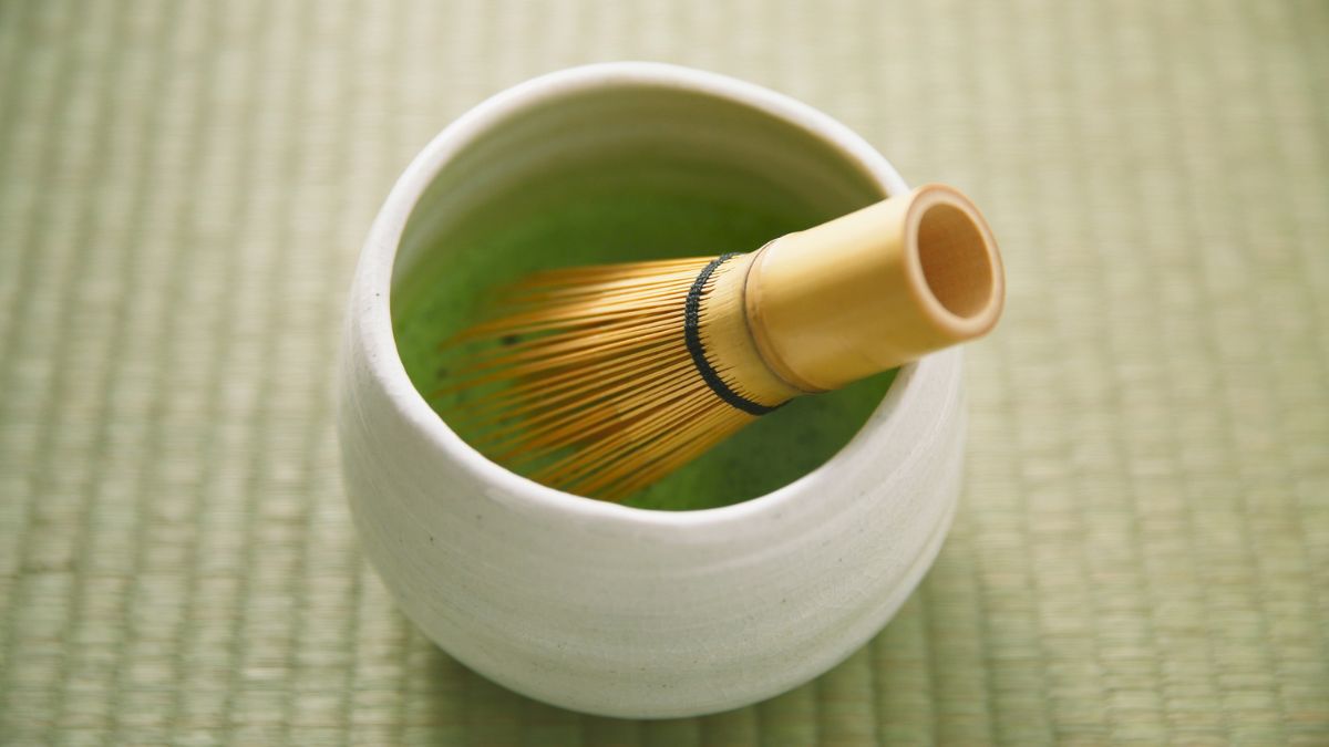 Miska s matcha čajom a bambusovým metličkom na zelenom podklade.