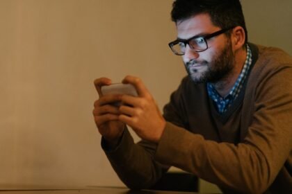 Muž sedí pri stole a sleduje obrazovku mobilného telefónu v tme.