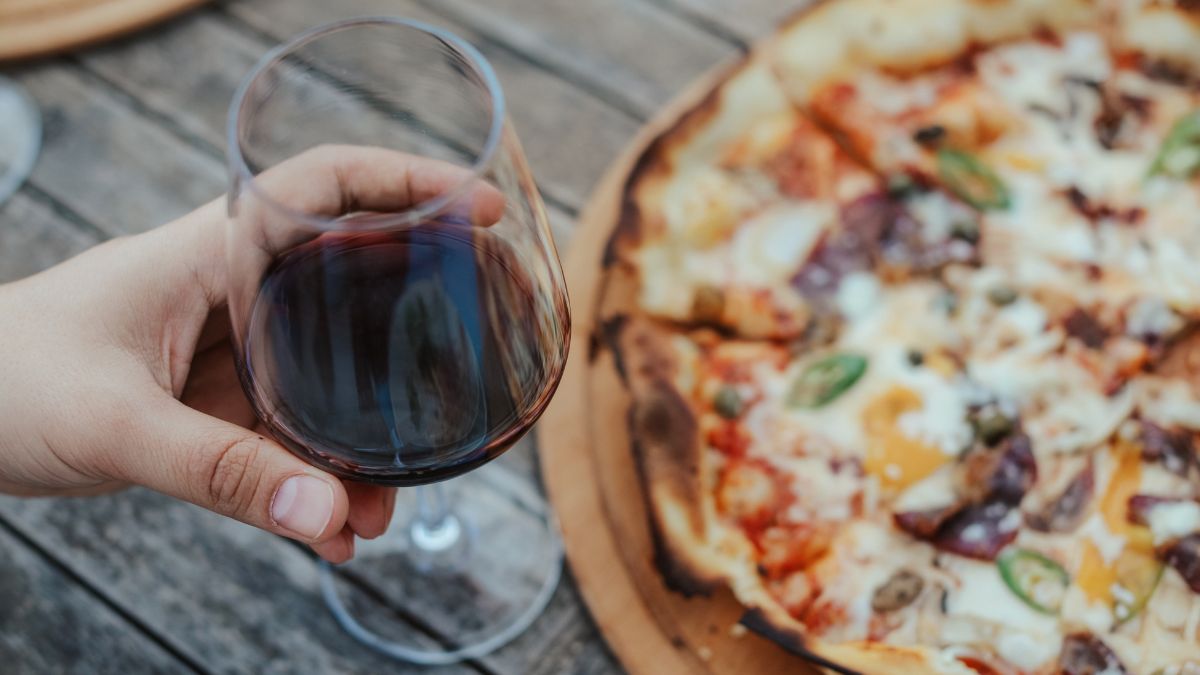 Ruka drží pohár červeného vína vedľa taniera s pizzou na drevenom stole.