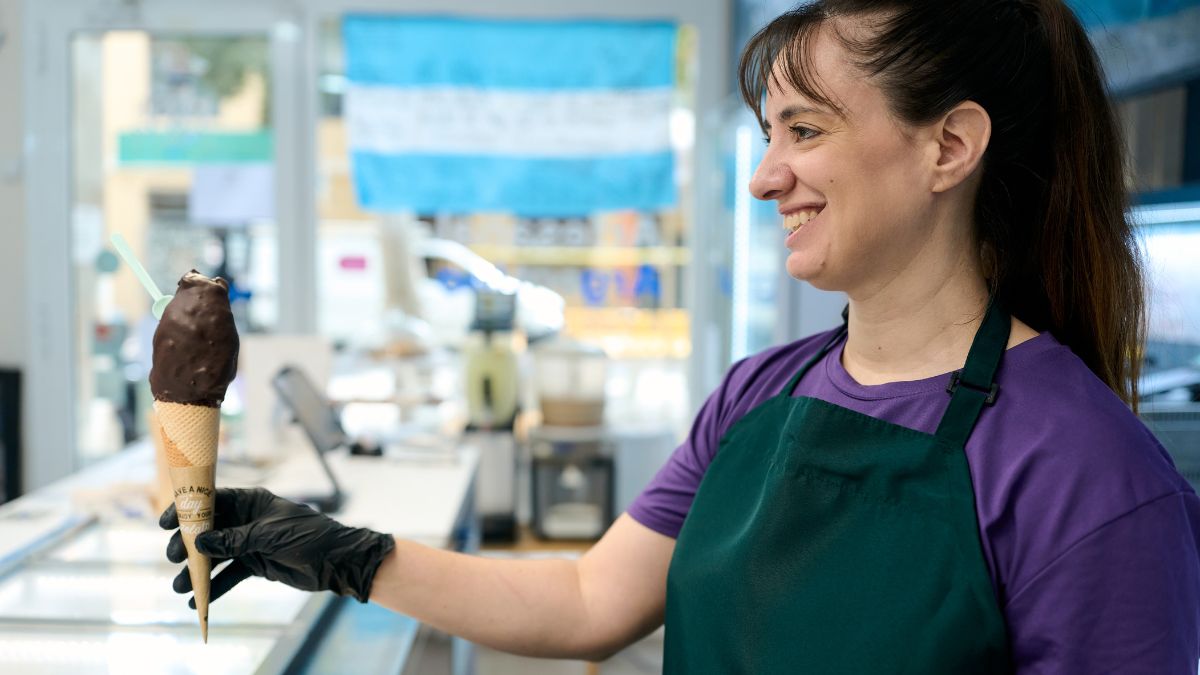 Žena s úsmevom drží zmrzlinu v kornúte, pozadie s modrou vlajkou.