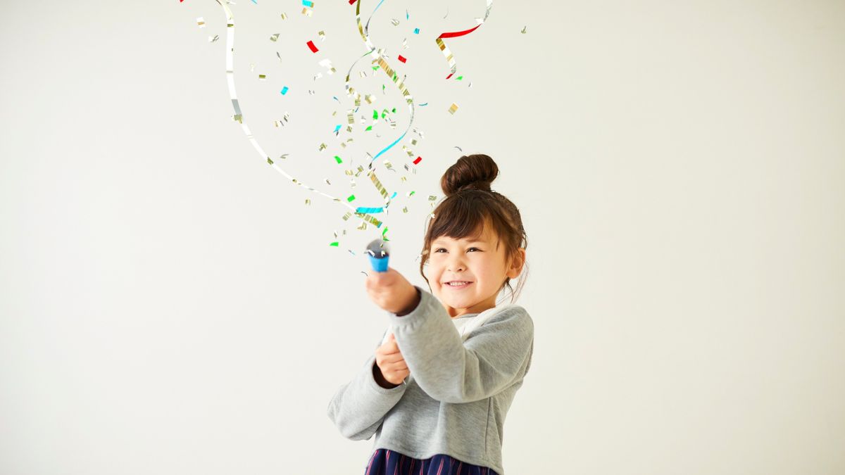 Dievča sa usmieva a strieľa konfeti z konfeti dela, okolo sú farebné kúsky.