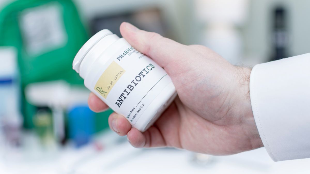 Ruka drží fľašu antibiotík, pozadie s lekárenskými potrebami.