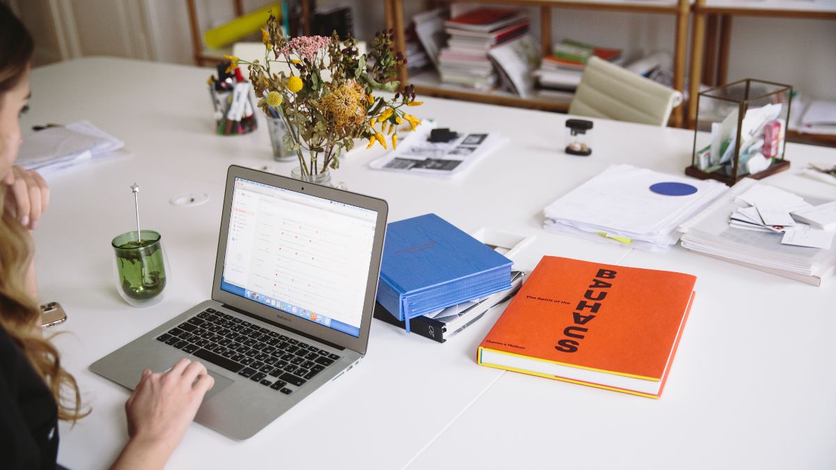 Žena pracuje na laptopu, okolo sú dokumenty a kvetinová dekorácia.
