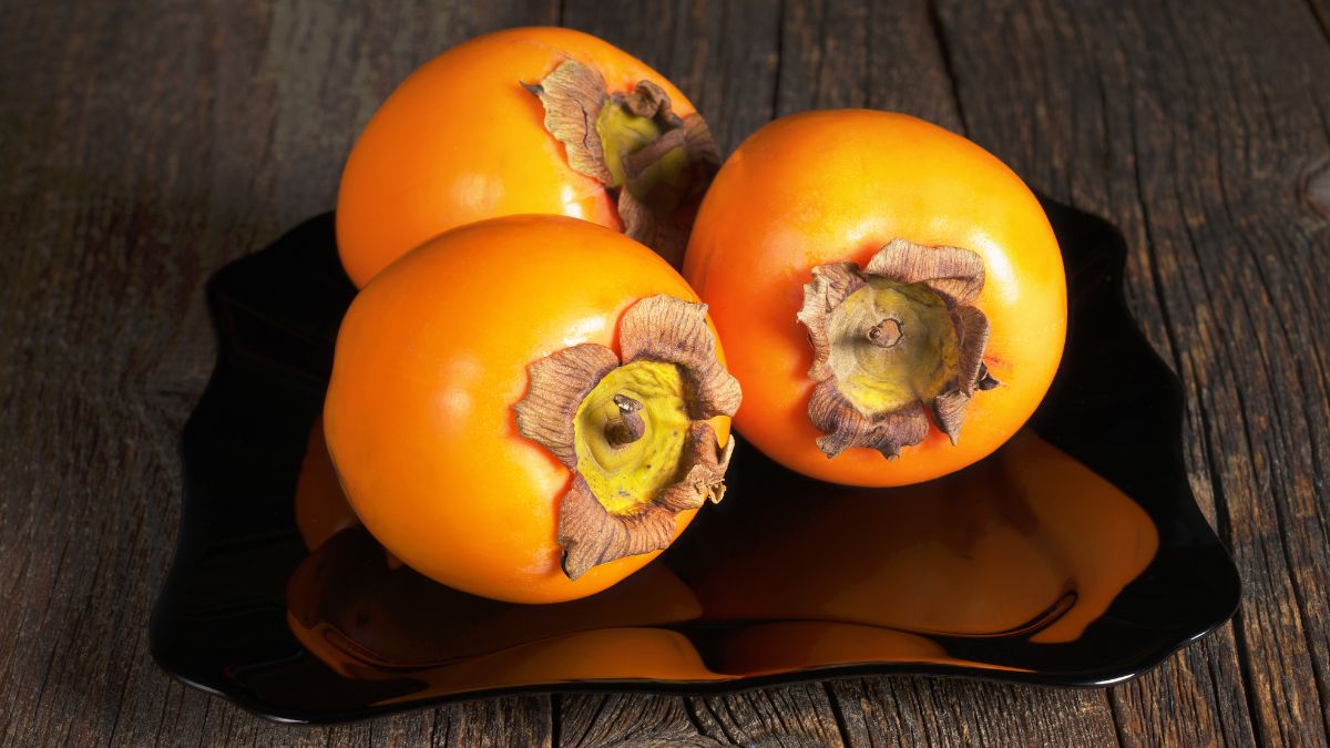 Tri zrelé kaki ovocie na čiernom tanieri na drevenom stole.