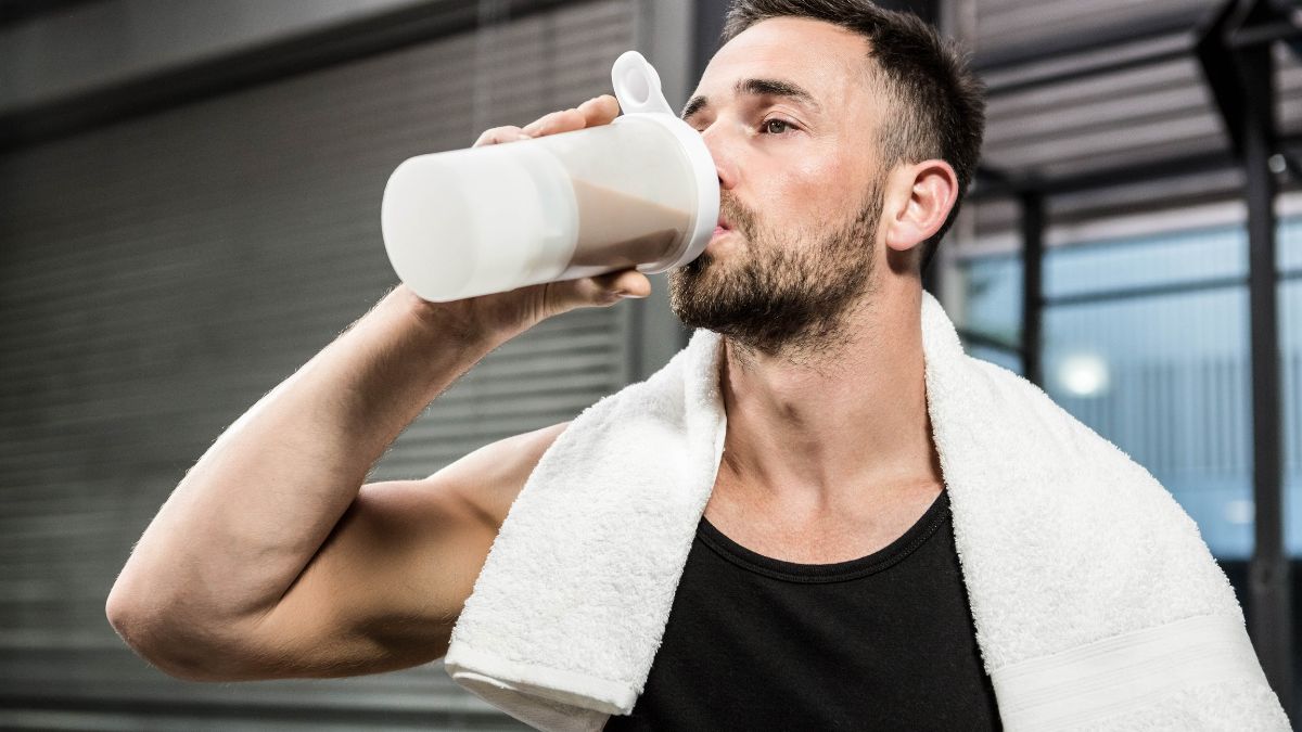 Muž v športovom oblečení pije proteínový nápoj po tréningu, s uterákom na krku.