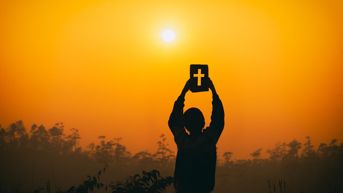 Osoba drží Bibliu s krížom proti slnku na pozadí oranžovej oblohy.