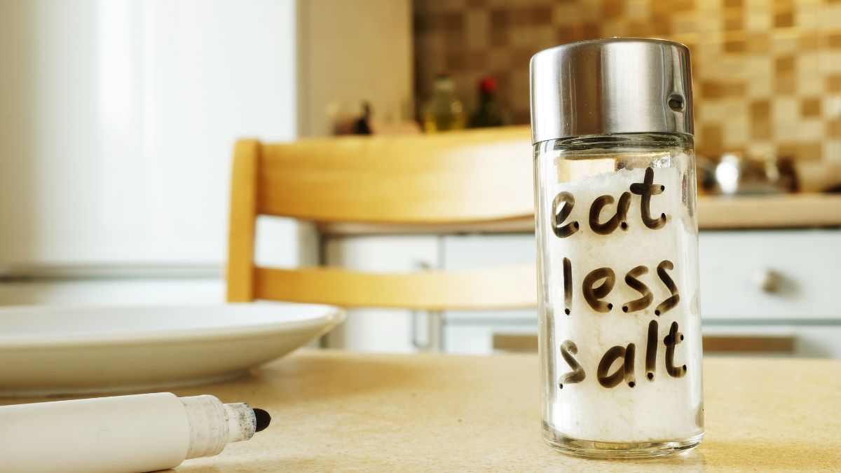 Sklenená nádoba so soľou s nápisom 'eat less salt' na kuchynskom stole.